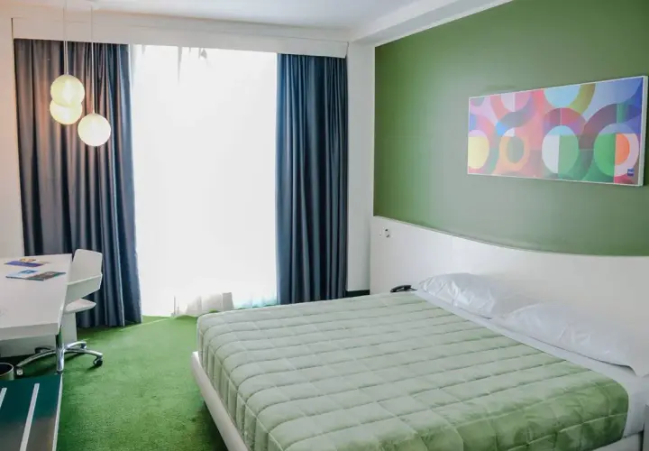 A modern hotel room with a green bedspread, green carpet, and a colorful abstract art piece on a green wall.