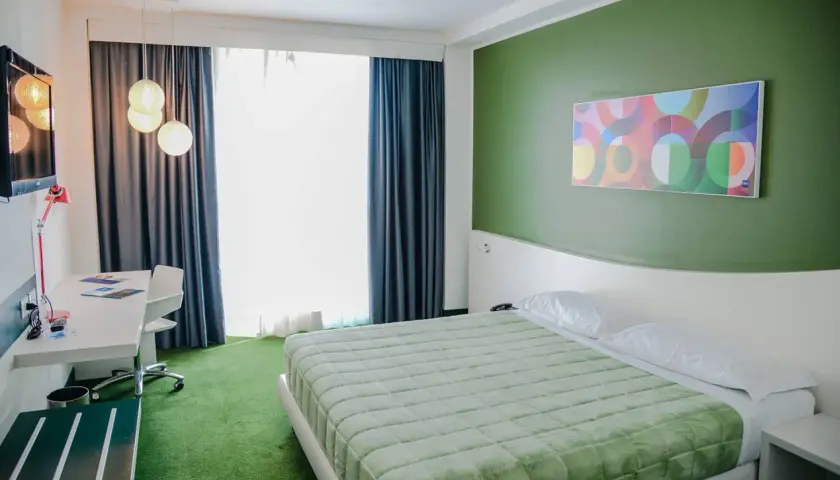 A modern hotel room with a green bedspread, green carpet, and a colorful abstract art piece on a green wall.